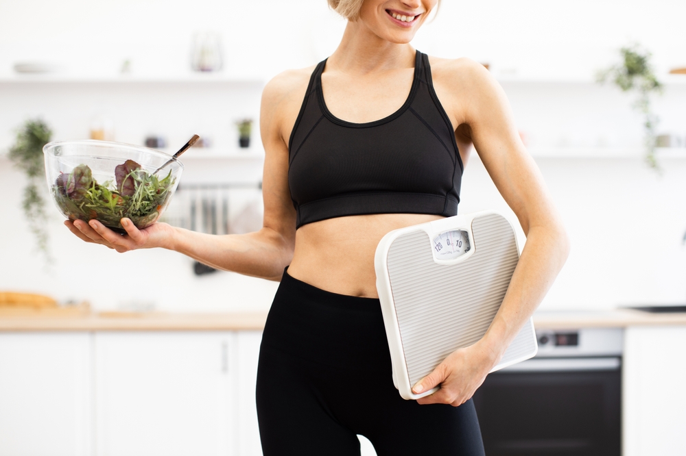 Fit woman in athletic wear holding salad bowl and bathroom scale in modern kitchen