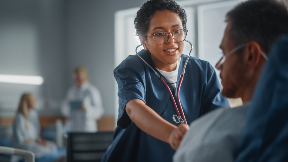 Friendly Black Head Nurse Uses Stethoscope to Listen to Heartbeat and Lungs of Recovering Male Patient Restin