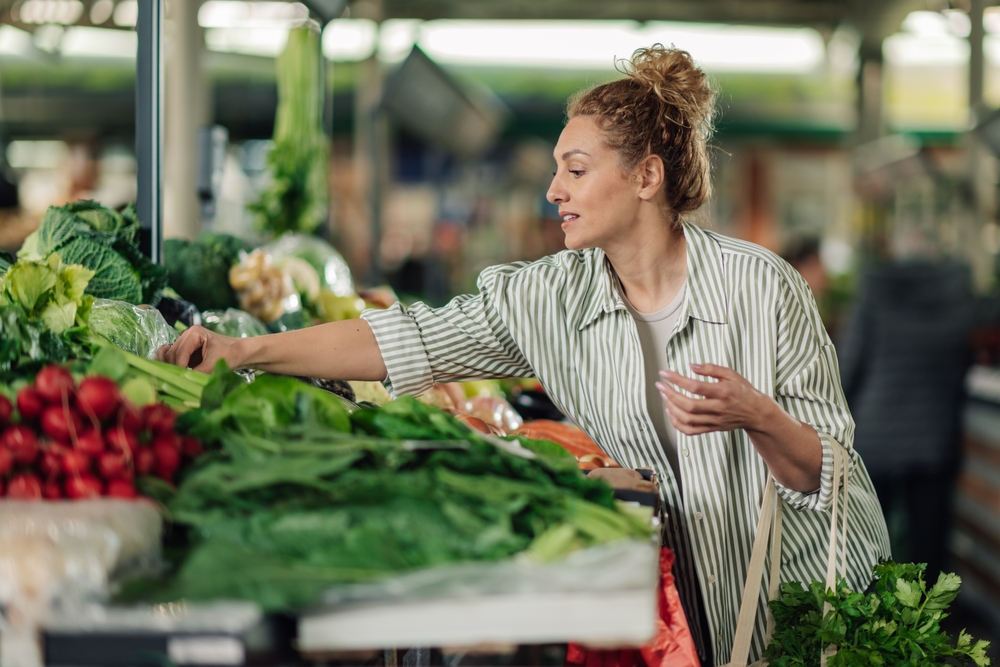 Portrait of smiling female customer standing near market stall at farmer's market and buying fresh organic vegetables