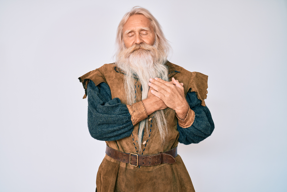 Old senior man with grey hair and long beard wearing viking traditional costume smiling with hands on chest