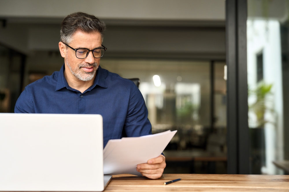 Busy middle aged professional business man using laptop computer reading document at work
