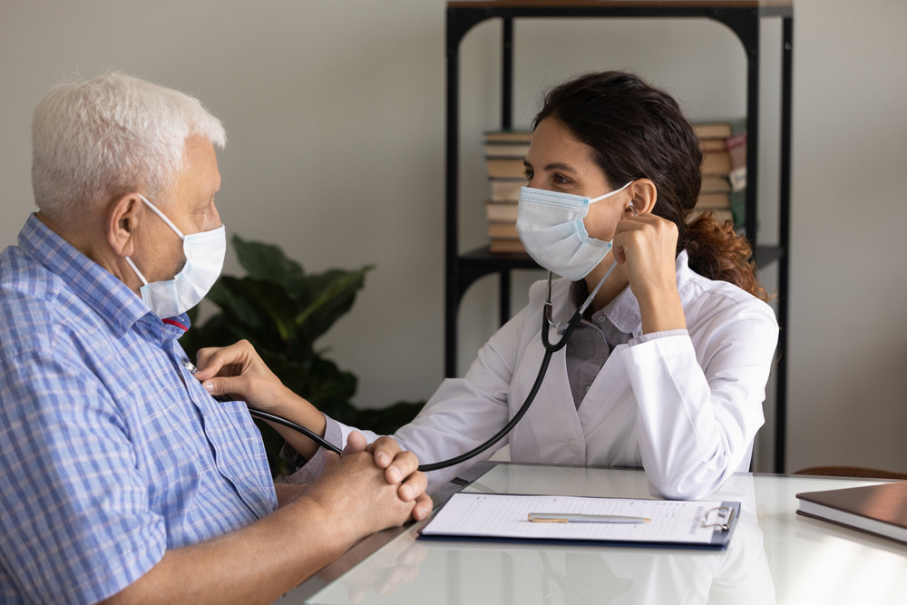 Caring female nurse in medical facemask listen to elderly patient heart with stethoscope in hospital
