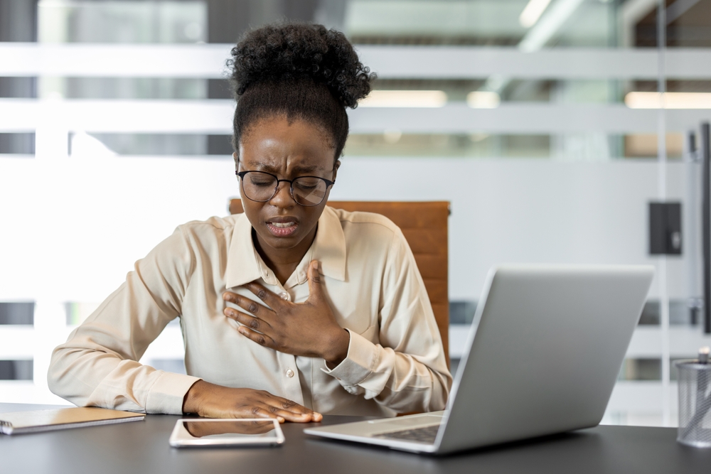 African American businesswoman sitting at office desk experiencing chest pain, possibly indicating heart attack or stress.