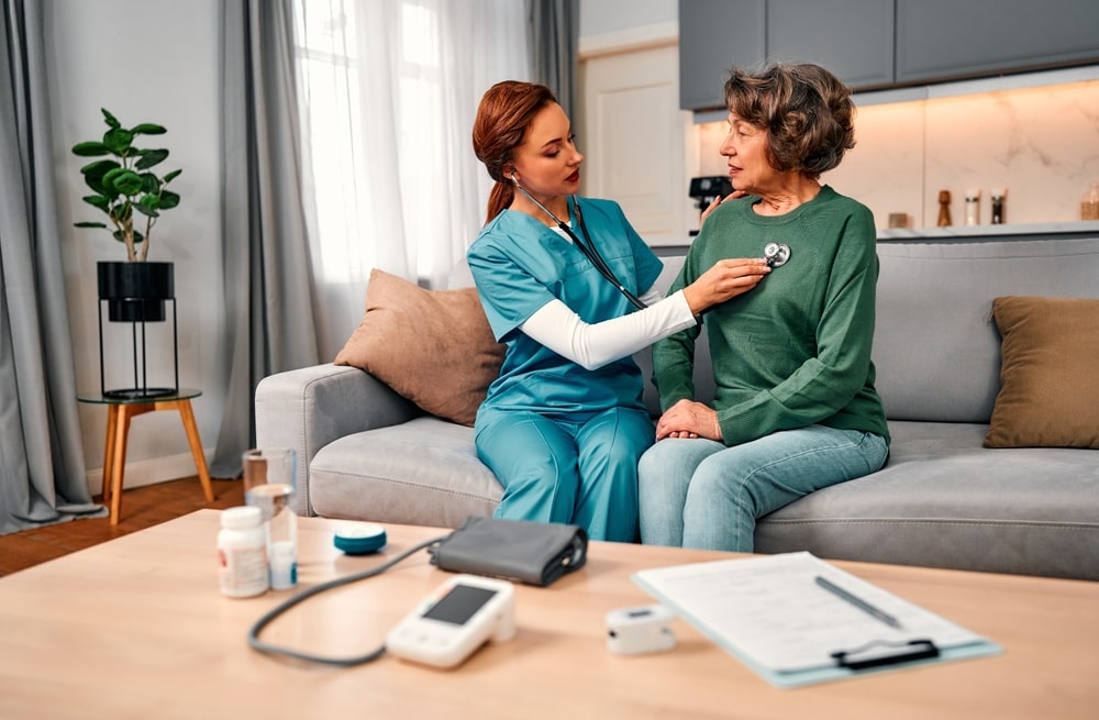 Doctor's home visit. Nurse in blue suit checking elderly woman with stethoscope while sitting on sofa at home. Health care,medicine.