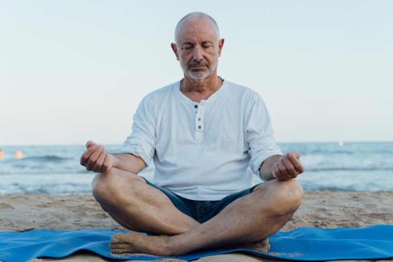 Older man meditating on the beach on a mat in the sand