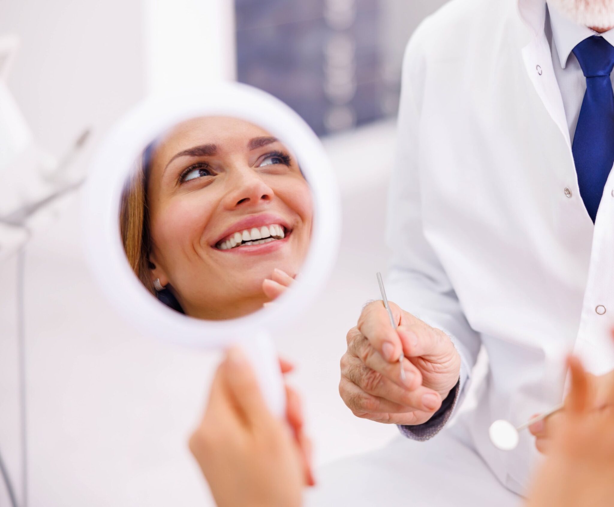 Woman looking in the mirror and smiling after checkup at dentist office; dentist and patient at dental clinic