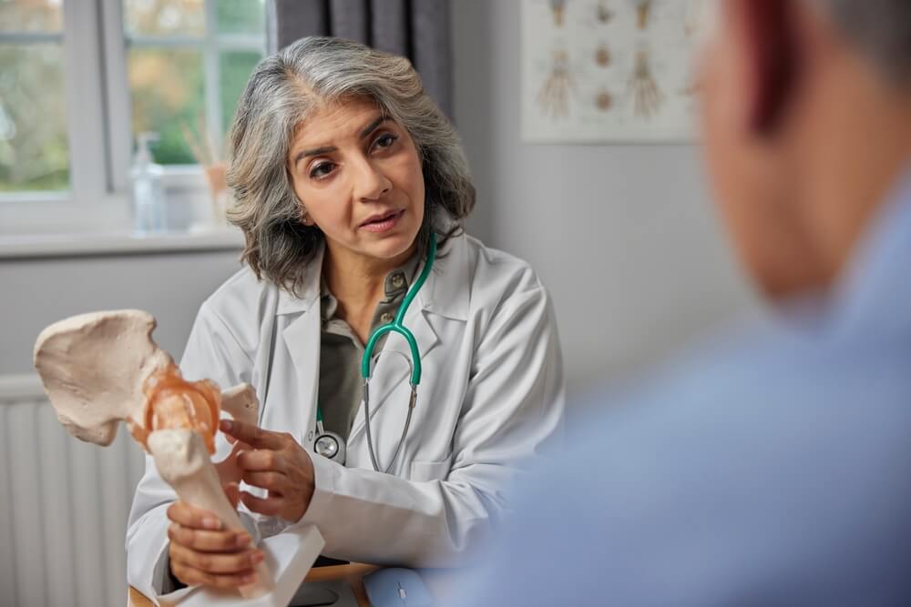 Mature Female Doctor Meeting With Male Patient Discussing Joint Pain In Hip Using Anatomical Model