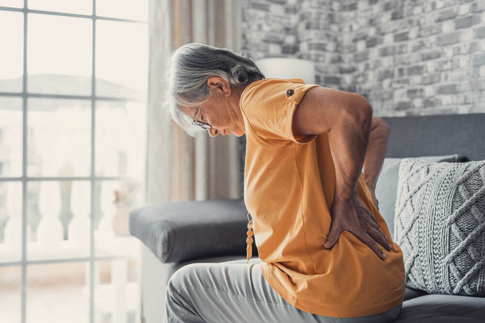 Unhappy mature woman touching back, feeling pain, sitting on couch in living room