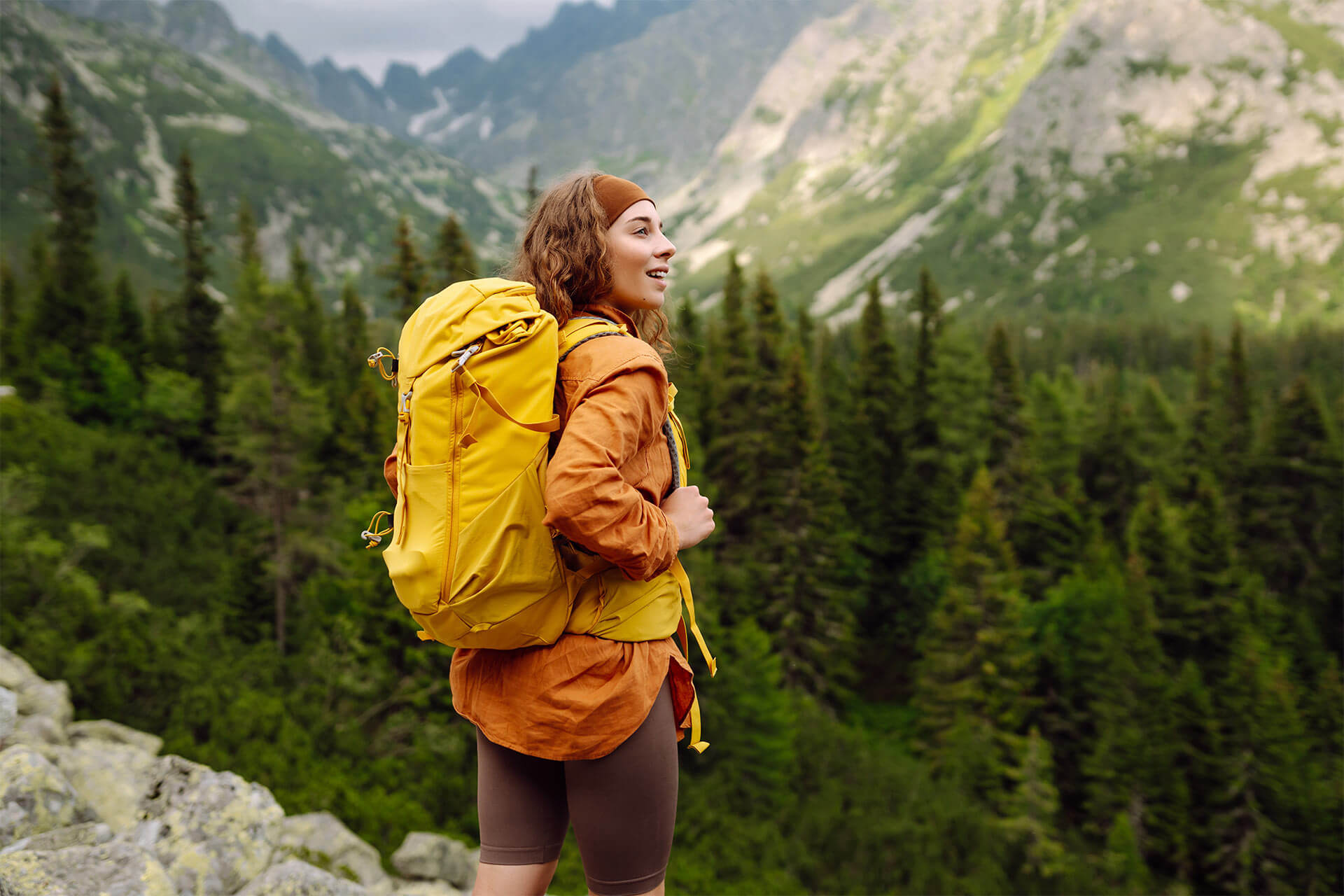 A smiling female traveler with a yellow hiking backpack enjoys the beautiful scenery of the majestic mountains