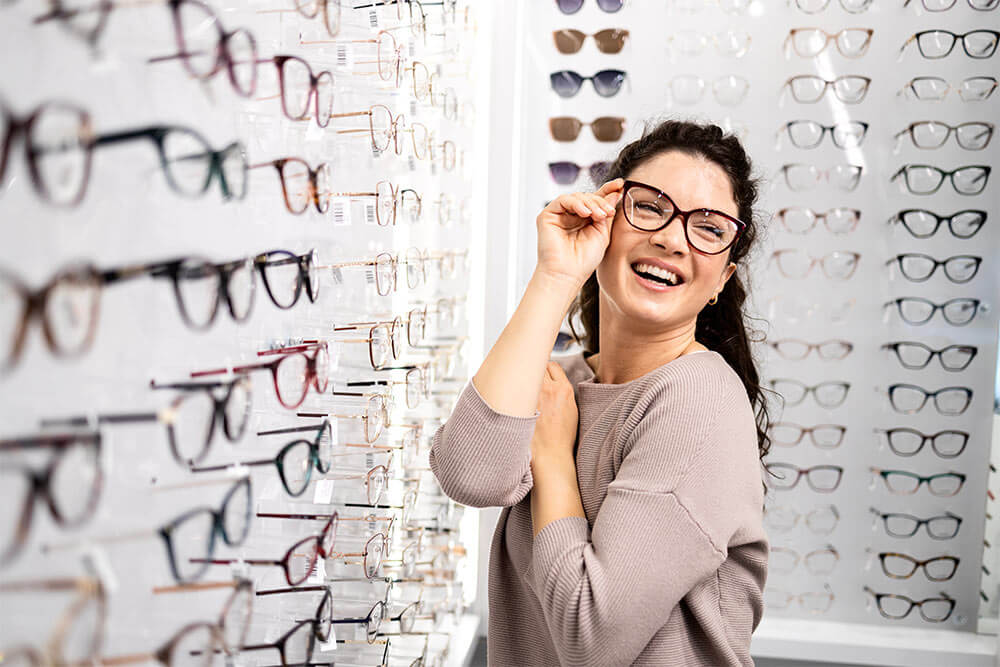 Beautiful smiling brunette woman trying on fashionable eyeglasses in optician store.