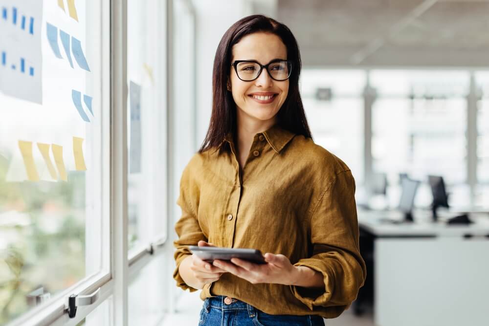 Thoughtful business woman holding a tablet while standing next to a window.