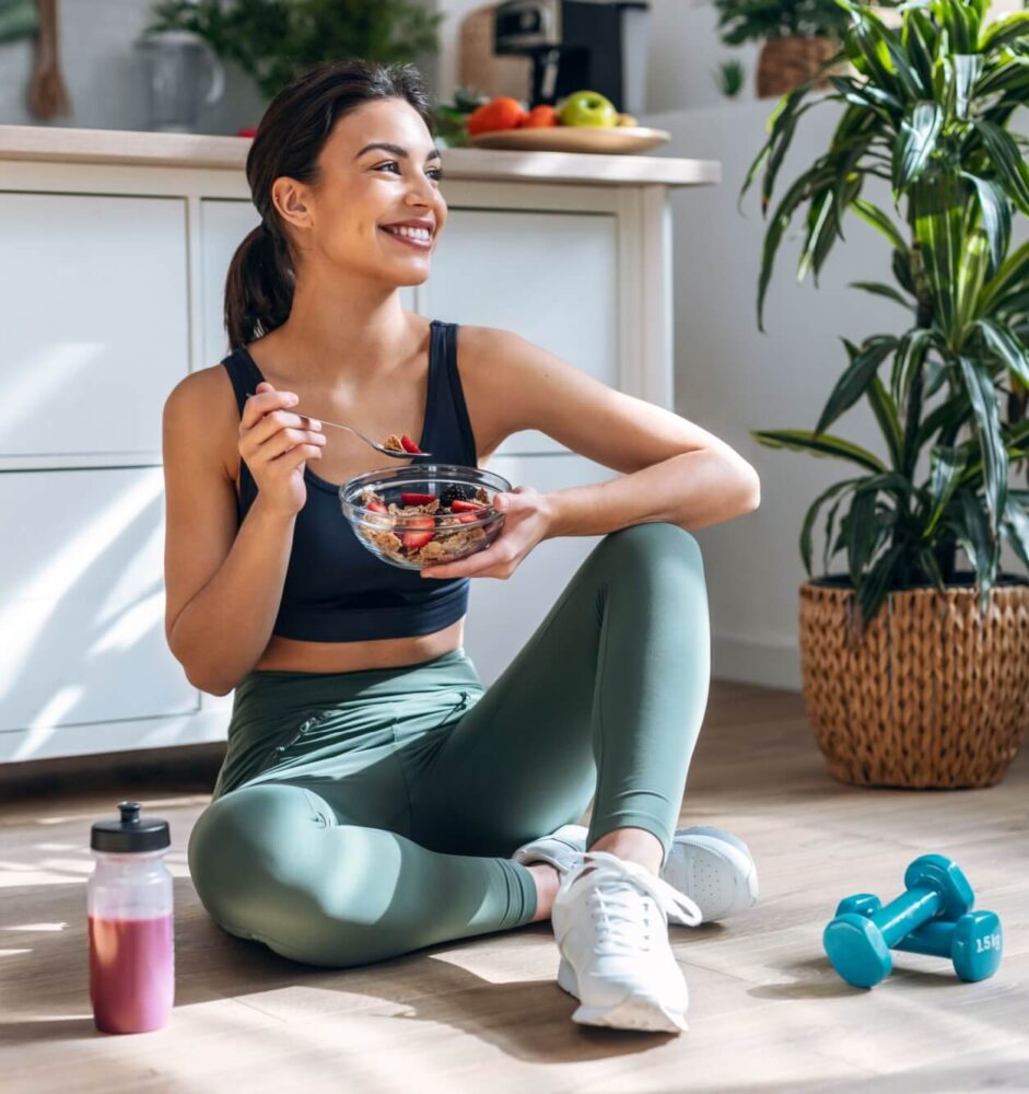 Shot of athletic woman eating a healthy bowl of muesli with fruit sitting on floor in the kitchen at home