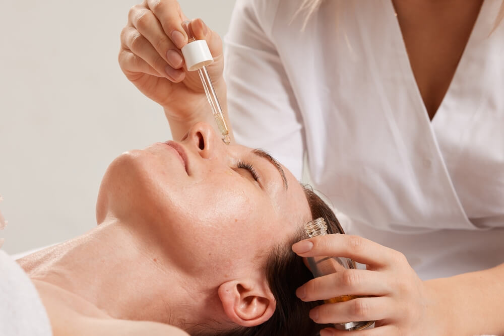 Cosmetologist applying facial oil serum with dropper to woman’s face during relaxing skincare spa treatment.