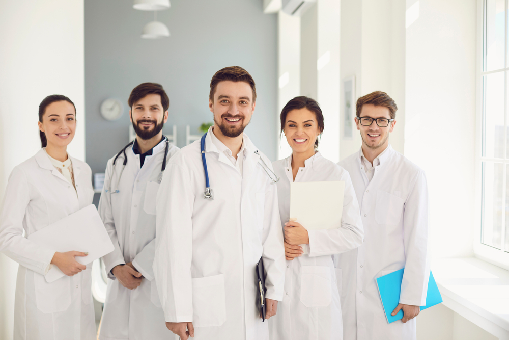 A group of confident practicing doctors in white coats are smiling against the backdrop of the clinic.