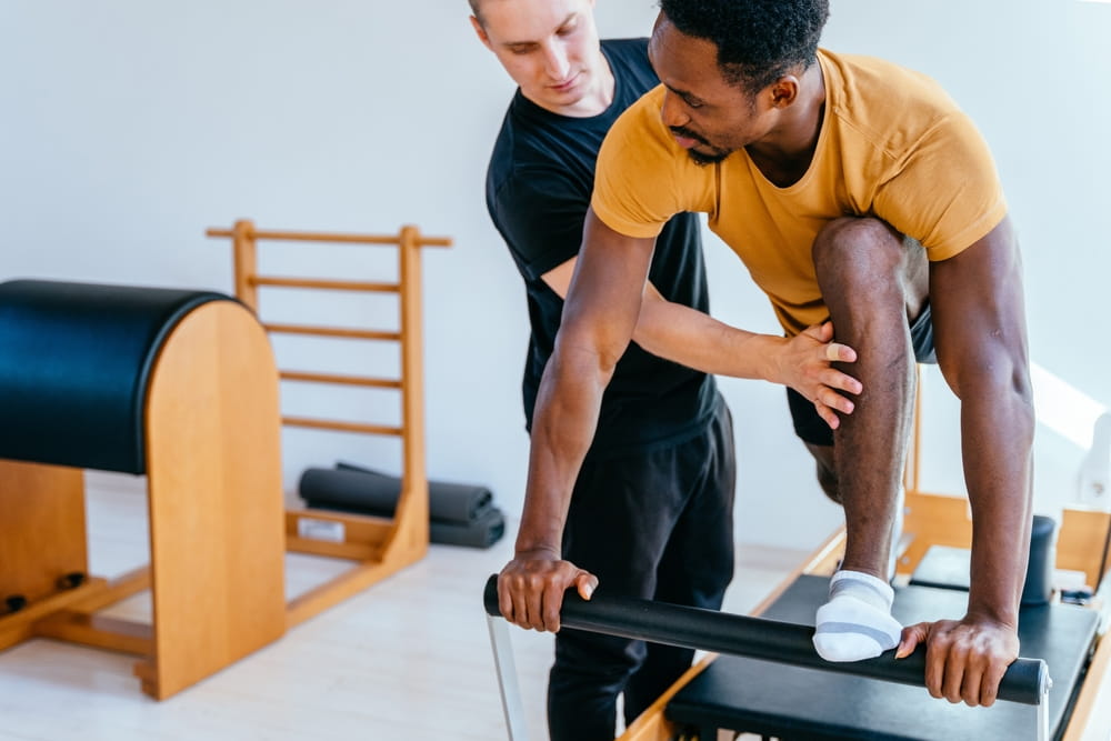 Fitness instructor corrects and controls the Pilates exercise that his african american male student is doing on Reformer bed