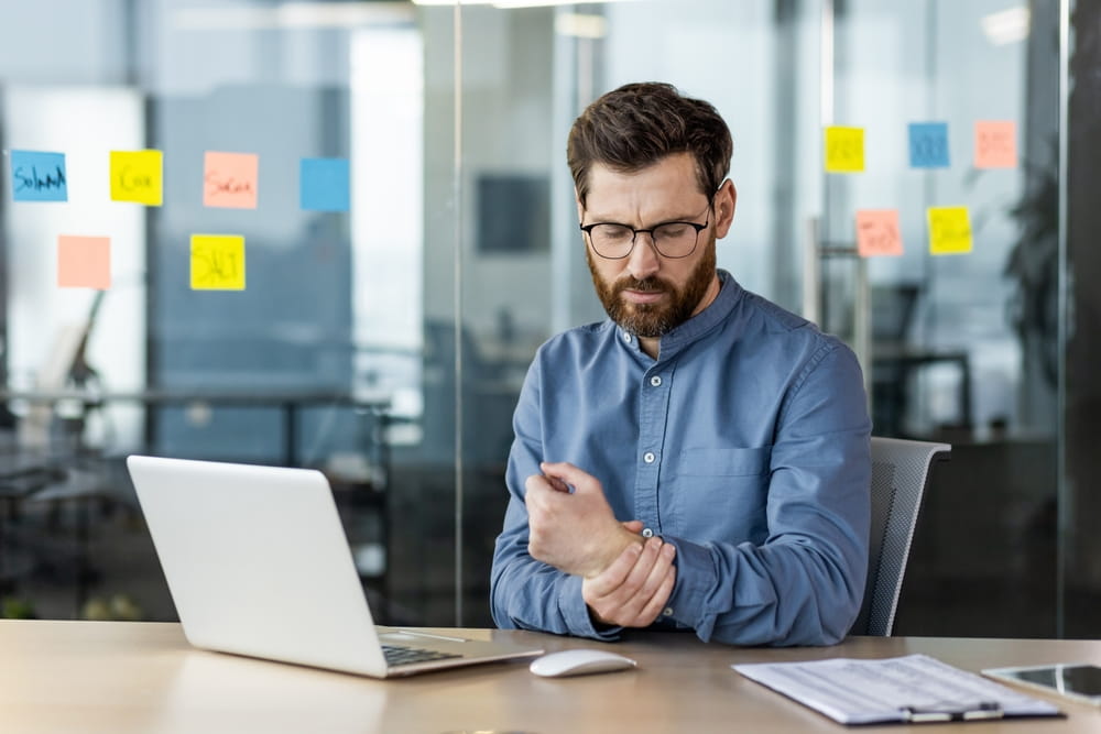 A tired young man works overtime in the office, sits at the table with a laptop, holds his hand by the wrist, does a massage, feels severe pain.