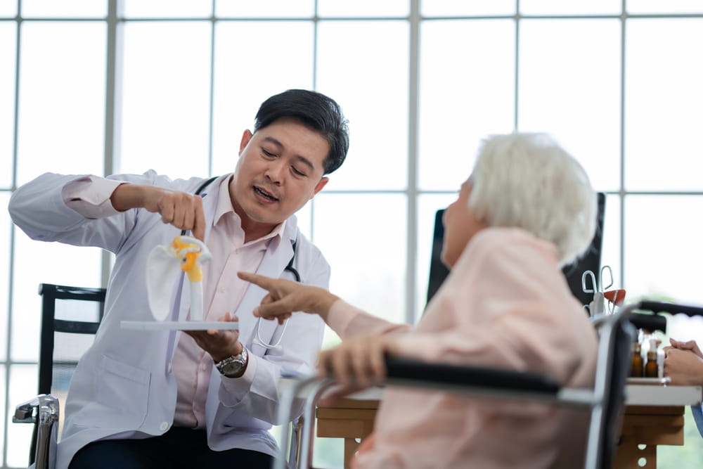 Orthopedic doctor consulting senior woman with joint injury and explaining treatment methods of trauma using anatomical model.