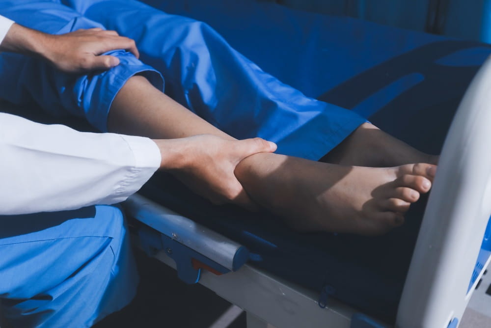 Asian male doctor examining foot of female patient sitting on bed in hospital room.