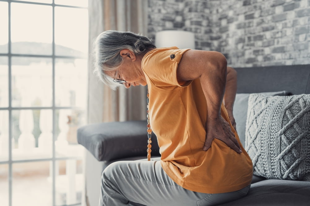 Unhappy mature woman touching back, feeling pain, sitting on couch in living room