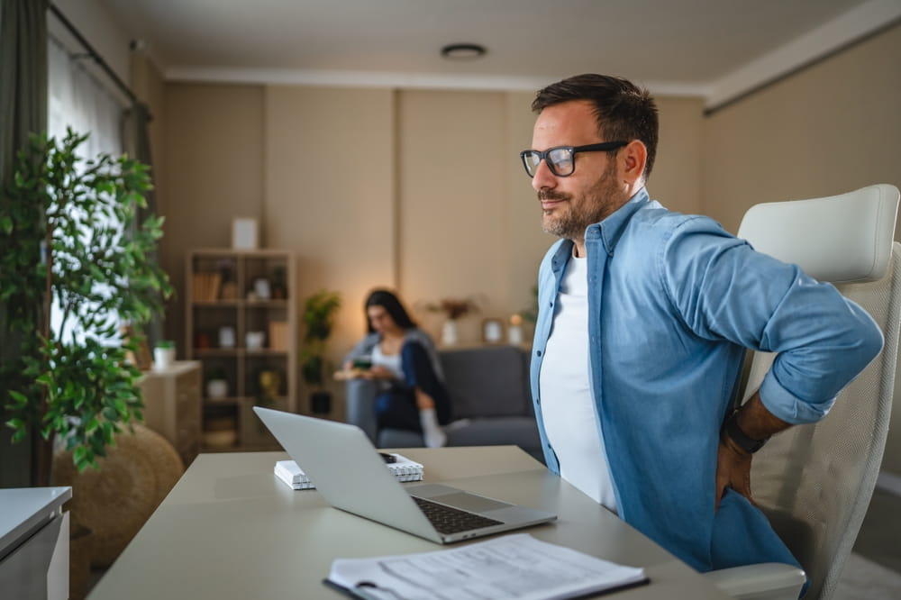 Man experiences discomfort and lower back pain while sitting at a desk and using a laptop in a home office