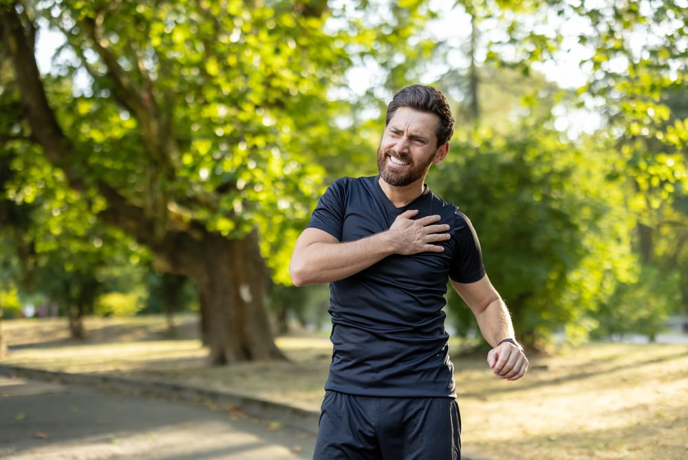A young male athlete, twisted in pain, stands in the park and holds his hand on his shoulder.