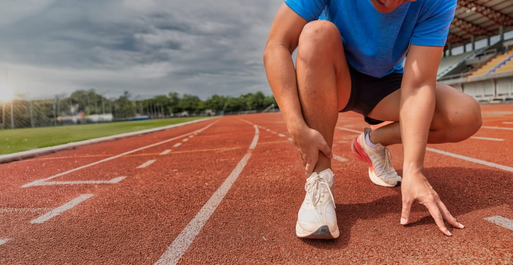 Athlete grabbing ankle in pain from injury or sprain on red running track field during sports activity