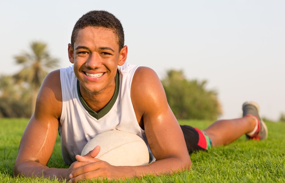 Portrait of happy fit African teen athlete relaxing on the grass with a rugby ball and smiling to camera.