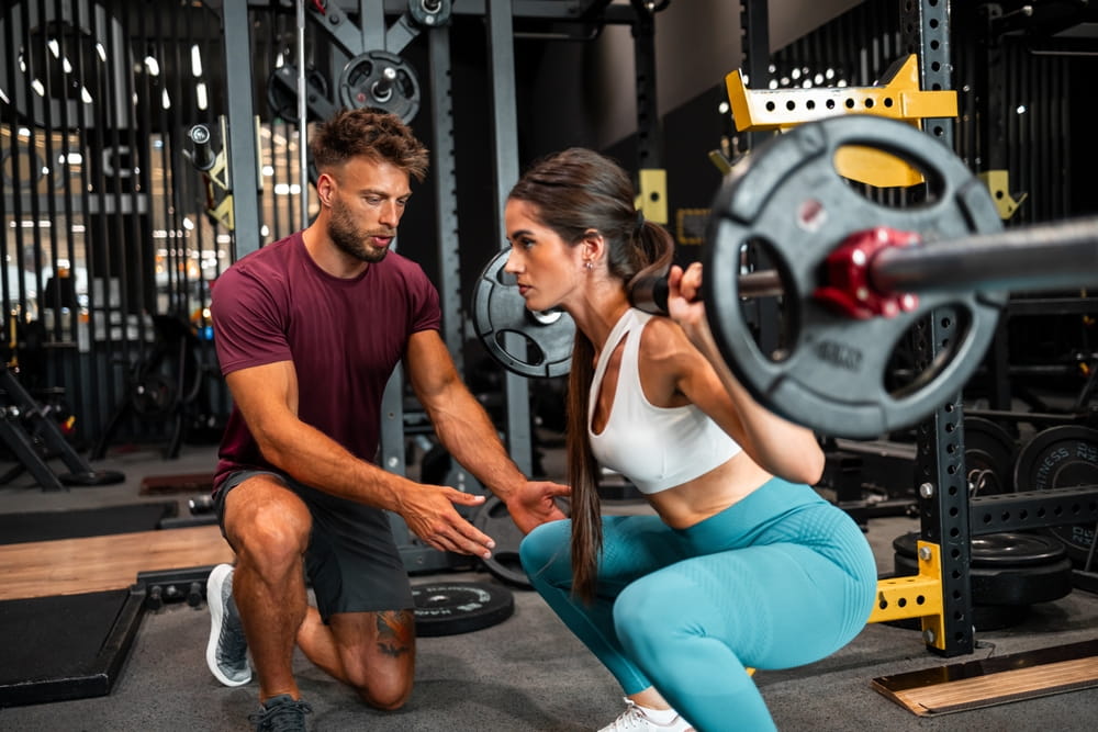 A trainer supervises a client's barbell squat, ensuring proper form and technique while exercising in a professional gym setting.
