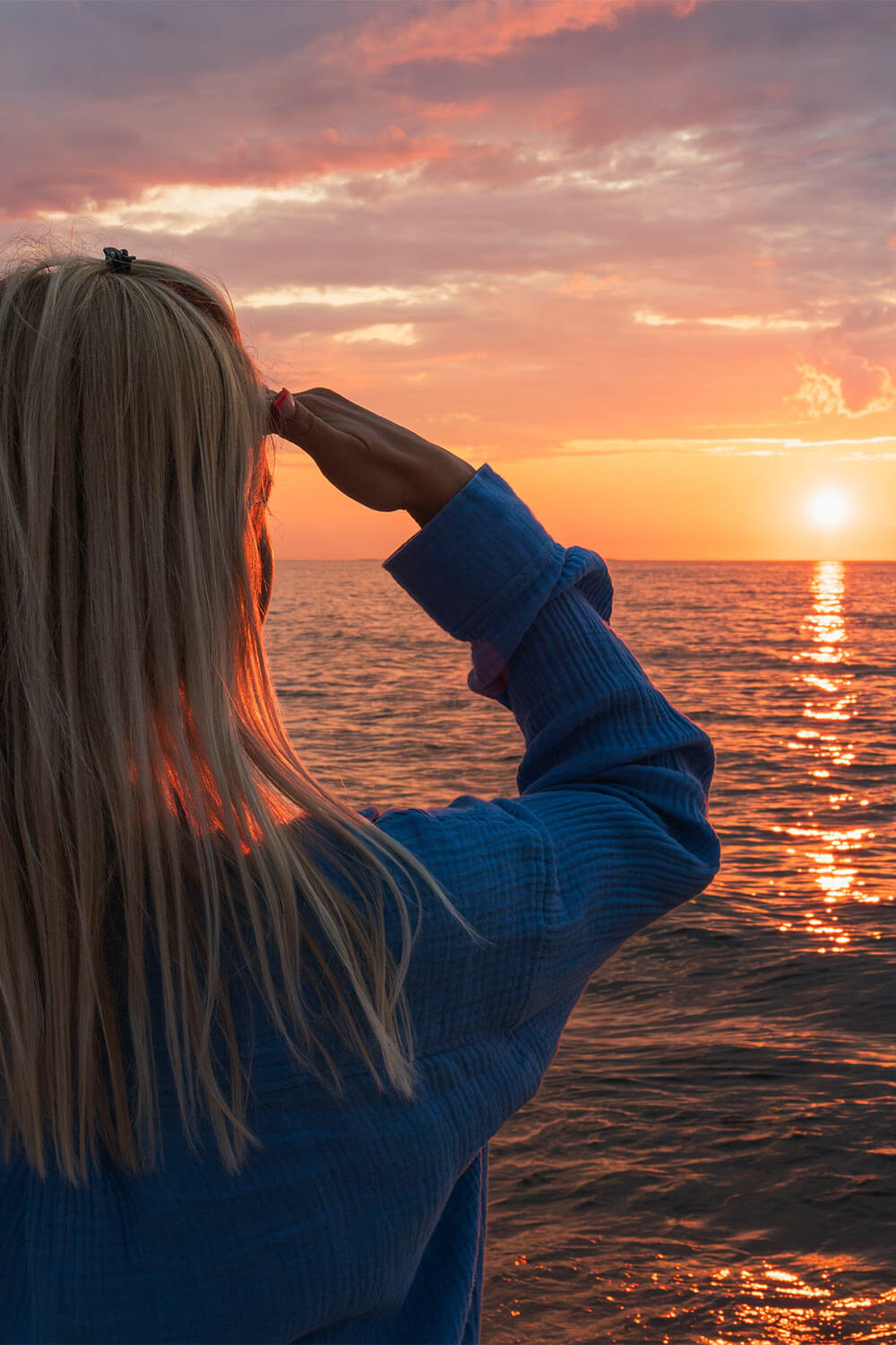 A dreamy blonde woman in a blue sweater enjoys the peaceful view of the sunset over the ocean