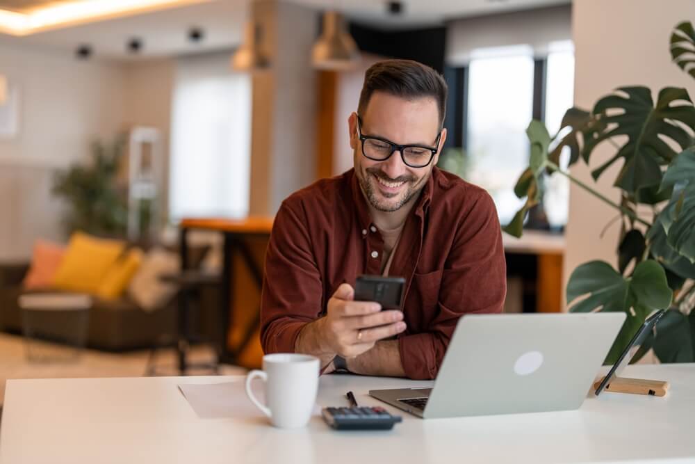 Happy smiling businessman wearing casual cloths and using modern smartphone in his home office