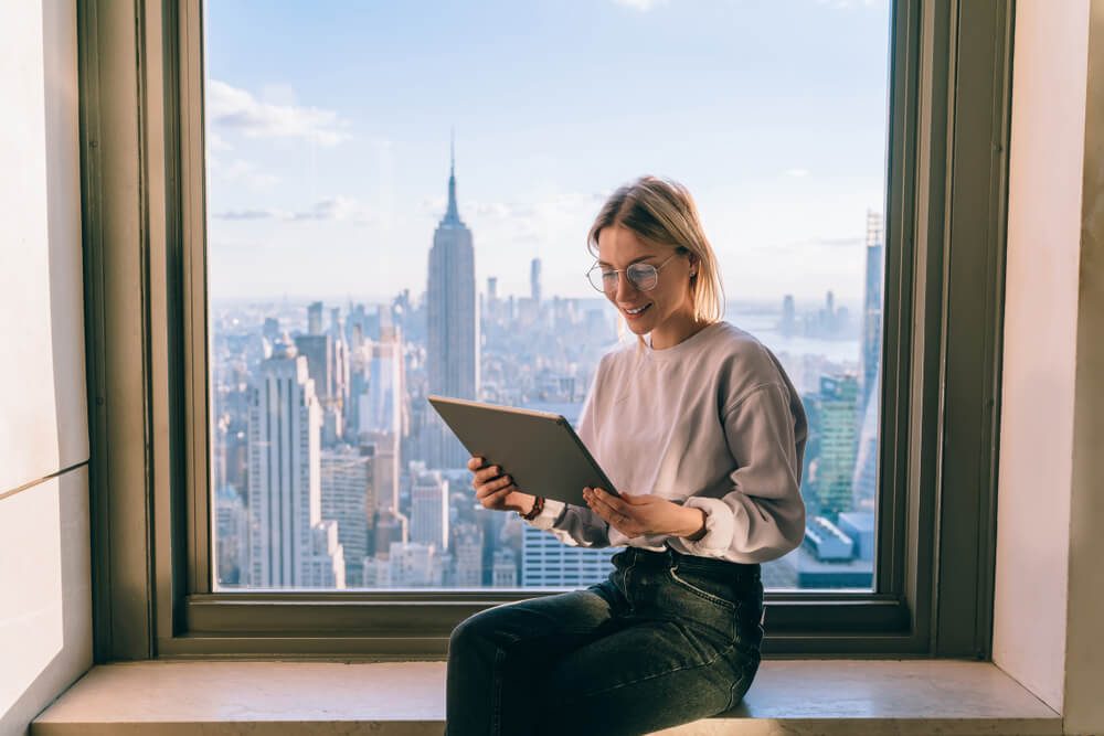 Smiling female office coworker working on modern tablet computer while sitting on window sill with panoramic views on New York city