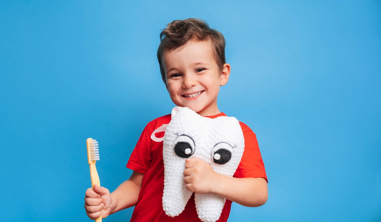 A smiling boy with healthy teeth holds a plush tooth and a toothbrush on a blue isolated background