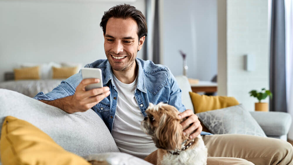 Young happy man text messaging on cell phone while sitting on the sofa with his dog.