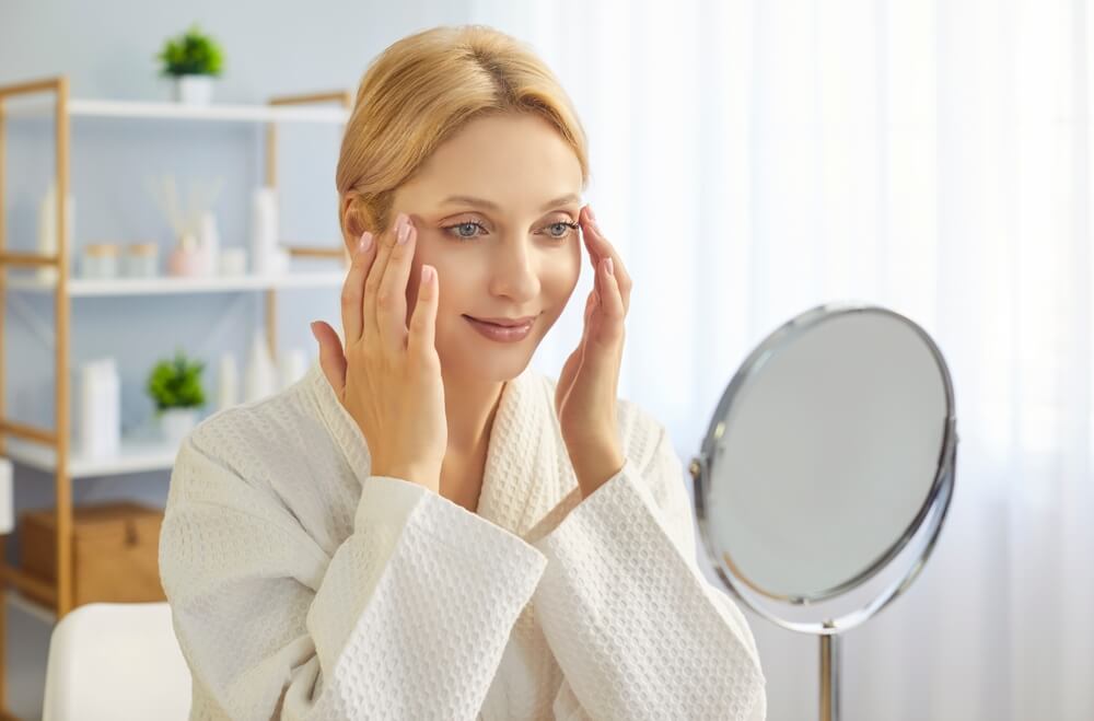 Young blonde woman in bathrobe gently applying skincare cream to her face using fingertips