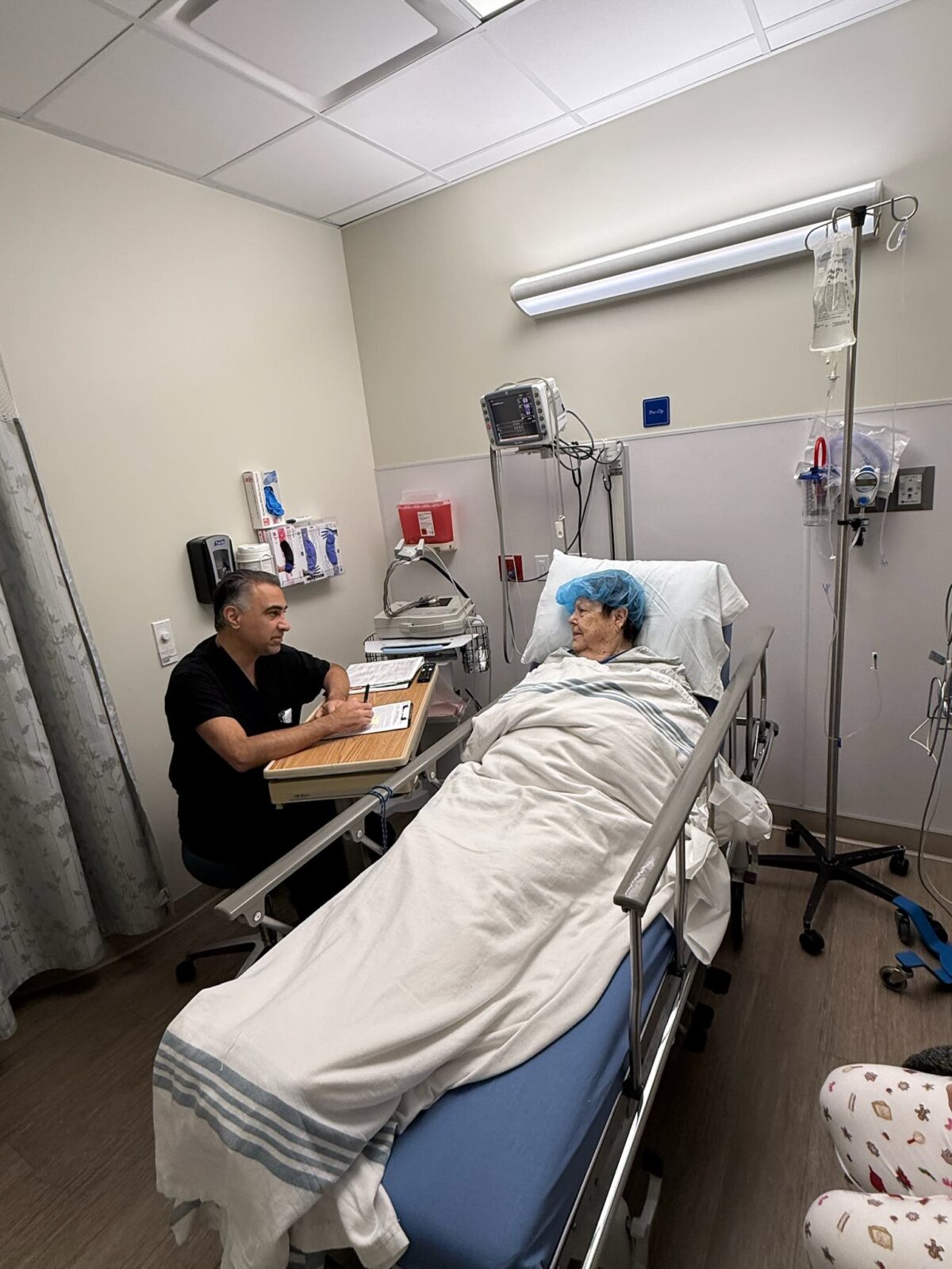 A medical professional in scrubs is seated nearby, likely reviewing charts or discussing the patient's care.