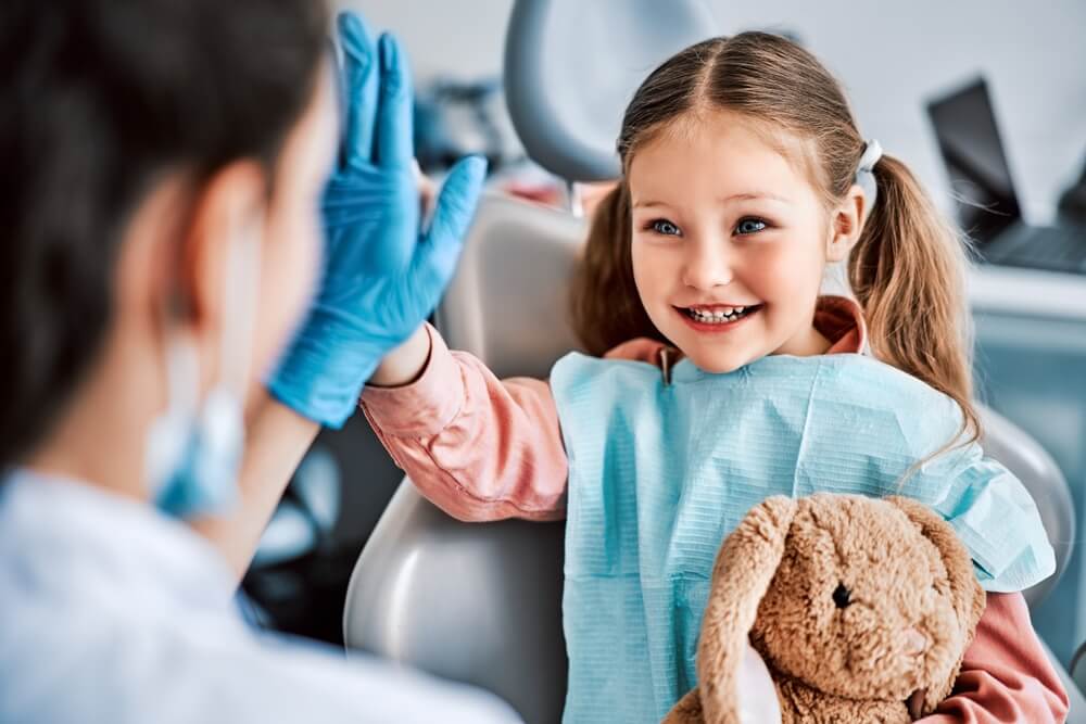 child sitting in a dental chair, holding a toy and cheerfully giving a high-five to the nurse