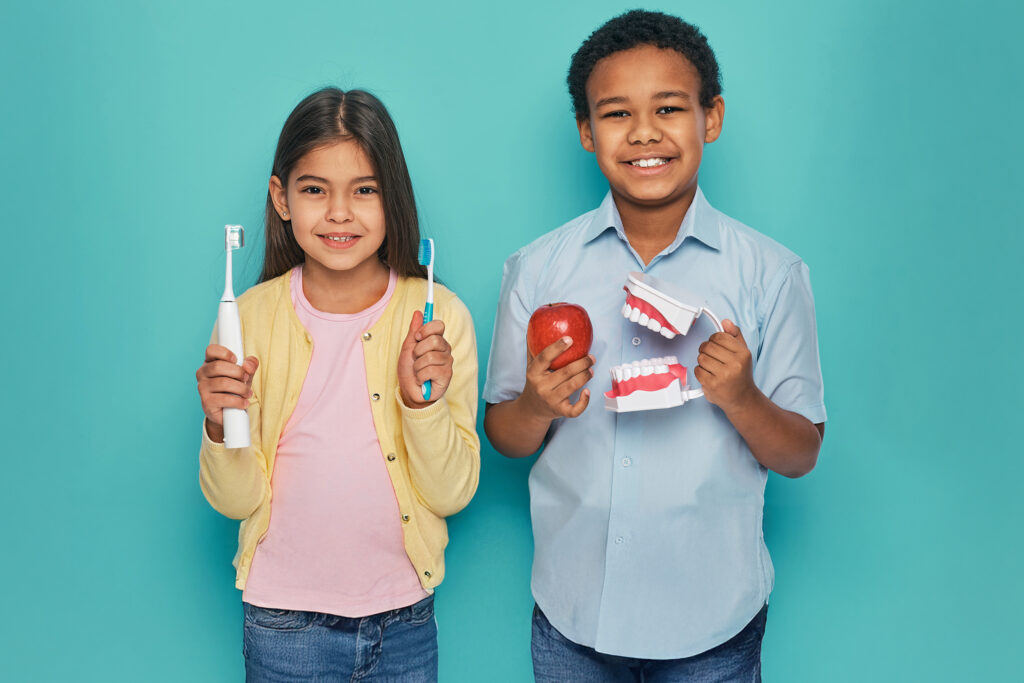 Girl is holding toothbrushes and boy is holding an apple and mouth model whikle smiling with teeth looking at camera