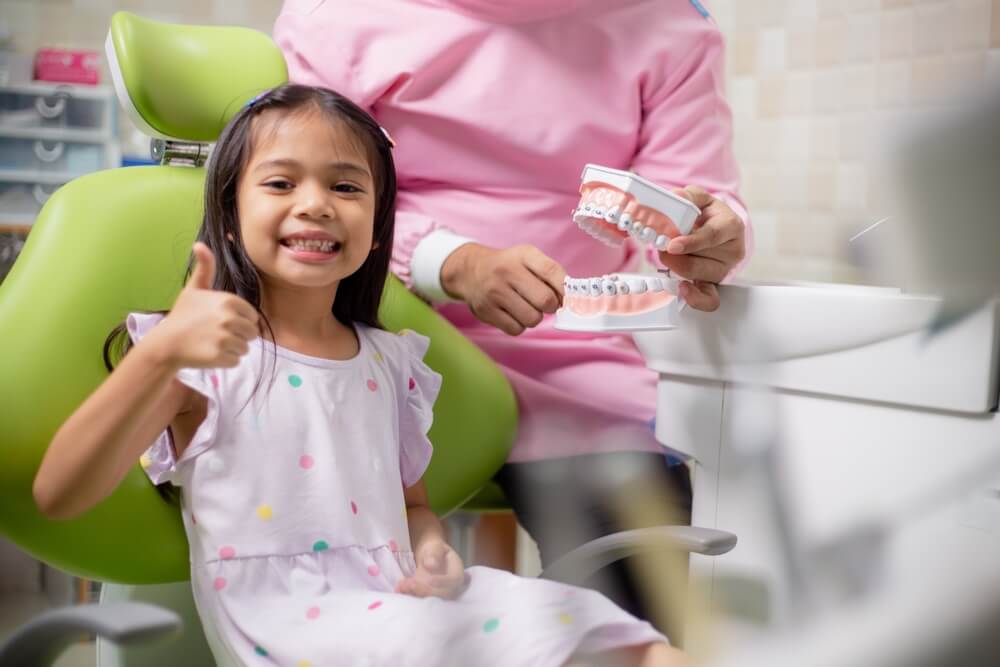 Young girl smiling showing teeth, sitting in a dental chair, and giving thumbs up next to a mouth model with braces that a dental assistant is holding