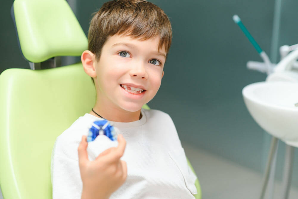 Young boy sitting in a dental chair smiling with teeth holding aligners