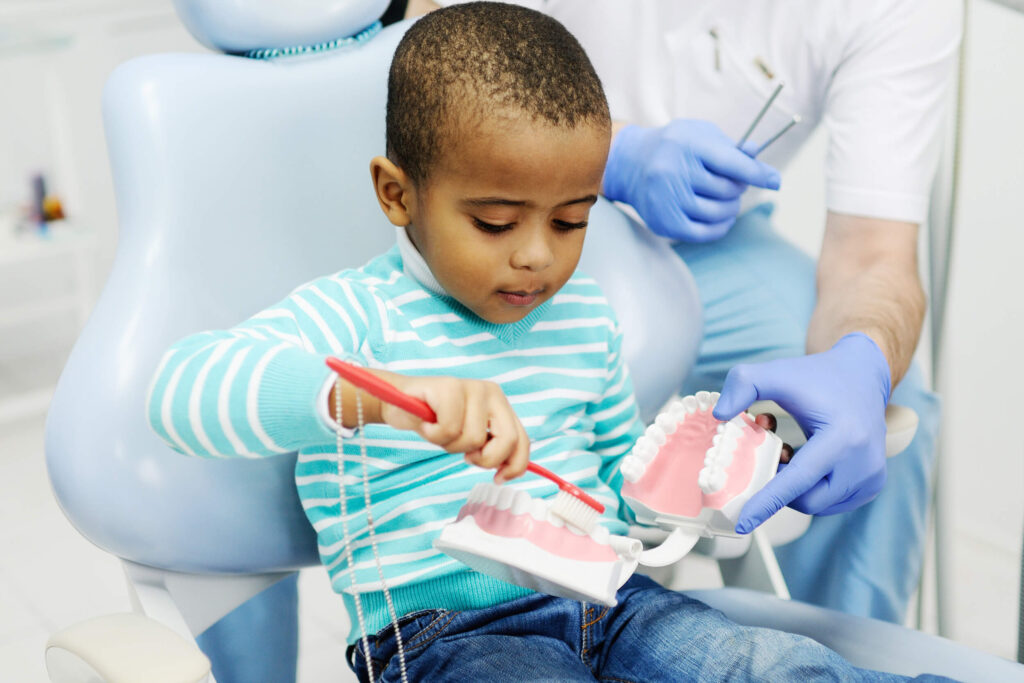 Toddler boy sitting in a dental chair and holding a red toothbrush and practicing brushing teeth on a mouth model that a dental assistant is holding