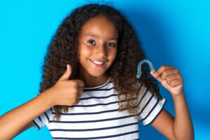 Young girl looking at camera and smiling with teeth while having a thumbs up and holding clear aligners