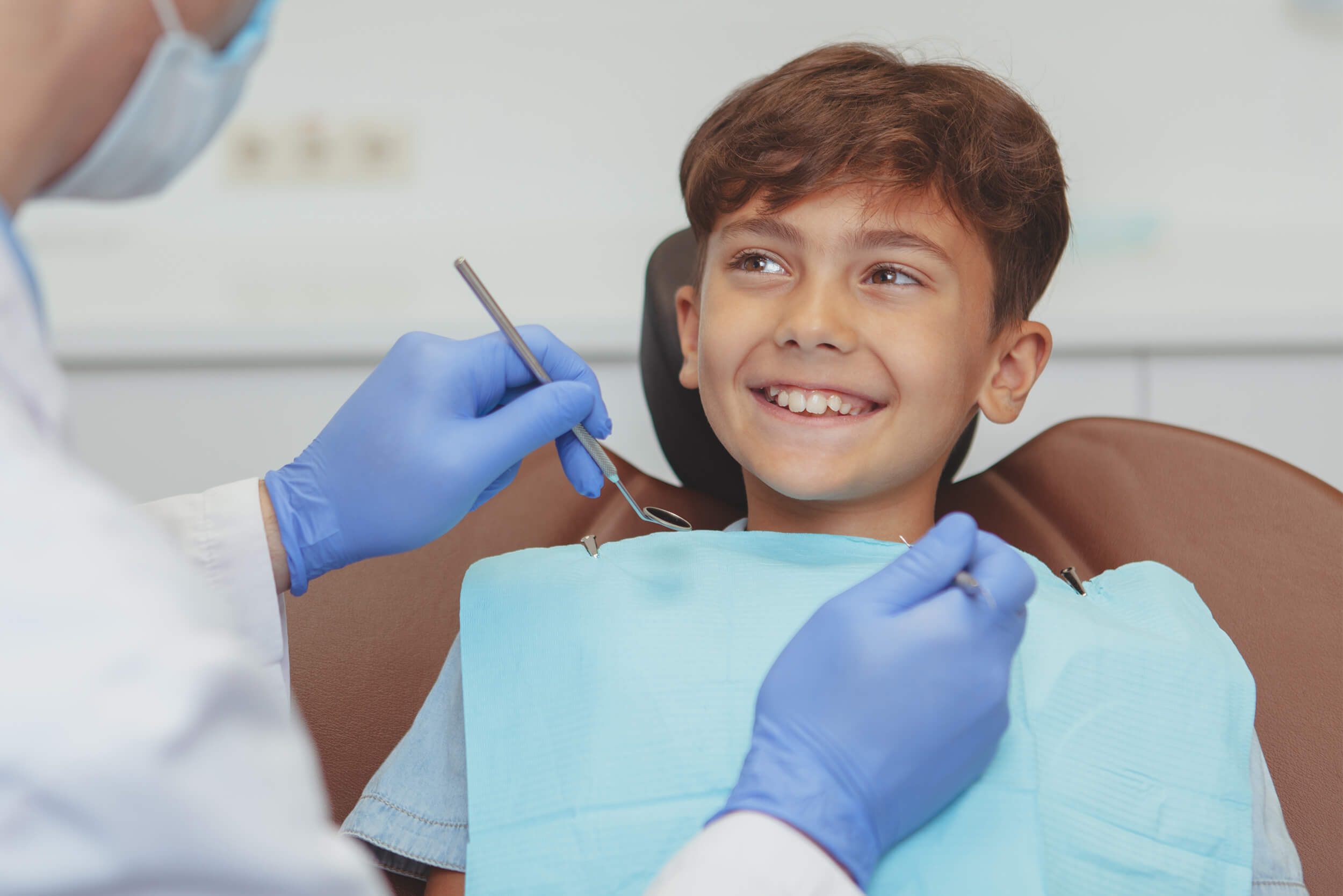 Young teenage boy sitting in a dental chair smiling with teeth while looking at dentist who is holding dental tools