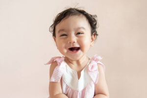 Girl toddler wearing a pink dress while smiling big with an open mouth showing baby teeth