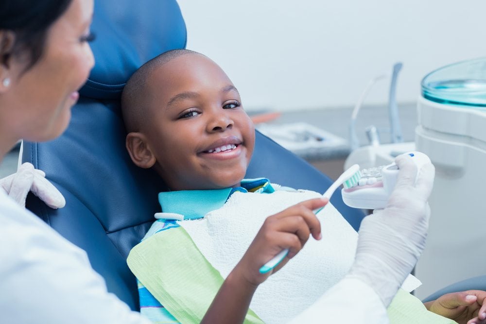 Young boy smiling while sitting in dental chair holding tooth brush ad practicing brushing teeth on mouth model that dental professional is holding