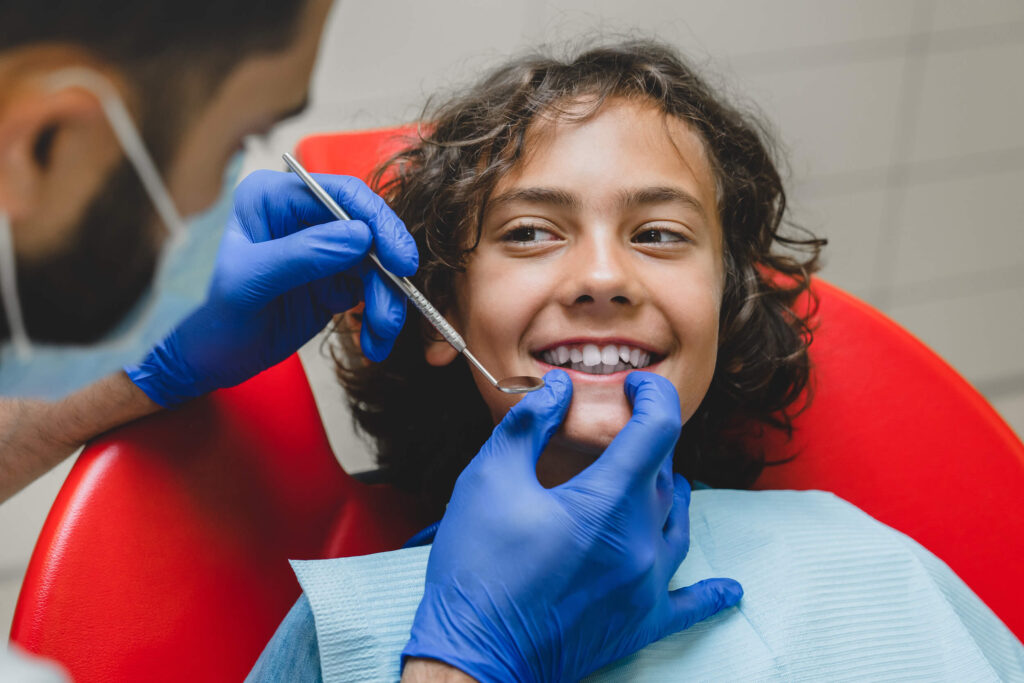 Young teenage boy sitting in a dental chair smiling with teeth while looking at dentist who is holding dental tools