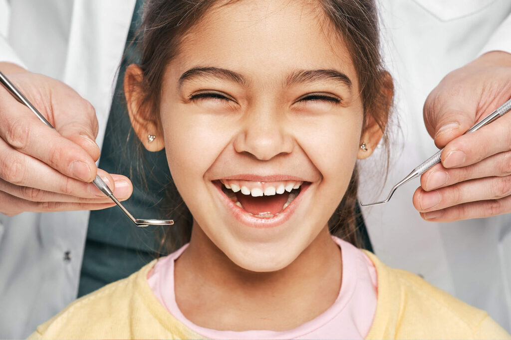 Young girl smiling and showing teeth about to get an exam. Dentist's hands are holding exam tools