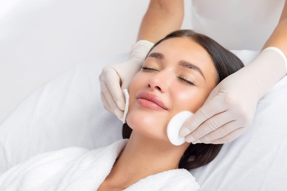 Female beautician cleansing skin of young woman